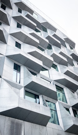 The image features a modern building facade with an intriguing geometric design. The structure is composed of numerous angular, protruding elements that cast subtle shadows. The facade is predominantly gray with glass windows interspersed among the geometric sections, reflecting a bit of light. The architectural style is contemporary and innovative, suggesting a focus on both aesthetics and functionality.