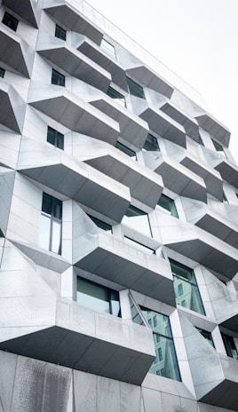 The image features a modern building facade with an intriguing geometric design. The structure is composed of numerous angular, protruding elements that cast subtle shadows. The facade is predominantly gray with glass windows interspersed among the geometric sections, reflecting a bit of light. The architectural style is contemporary and innovative, suggesting a focus on both aesthetics and functionality.