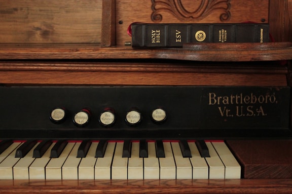 An antique organ with a series of labeled stops and white and black keys. The words 'Brattleboro, Vt., U.S.A.' are visible on the organ. A closed Holy Bible in the English Standard Version is resting on a wooden shelf above the keys.