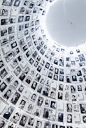 A large cylindrical wall filled with numerous black and white photographs, each accompanied by text. The images are historical portraits, arranged in a circular pattern that extends upwards, creating a powerful visual narrative. The architectural structure is illuminated from above, highlighting the photographs.