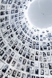 A large cylindrical wall filled with numerous black and white photographs, each accompanied by text. The images are historical portraits, arranged in a circular pattern that extends upwards, creating a powerful visual narrative. The architectural structure is illuminated from above, highlighting the photographs.