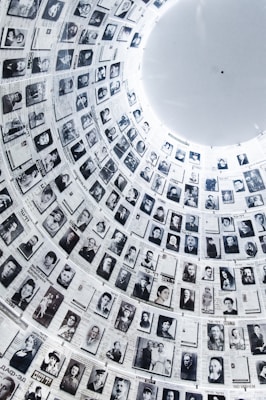 A large cylindrical wall filled with numerous black and white photographs, each accompanied by text. The images are historical portraits, arranged in a circular pattern that extends upwards, creating a powerful visual narrative. The architectural structure is illuminated from above, highlighting the photographs.