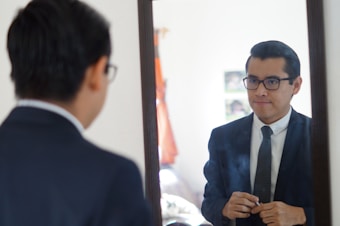 A man in a suit is gazing into a mirror, adjusting his clothing. He is wearing glasses and standing in what appears to be a softly lit room with some furniture and a partially visible window.