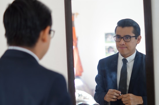 A man in a suit is gazing into a mirror, adjusting his clothing. He is wearing glasses and standing in what appears to be a softly lit room with some furniture and a partially visible window.
