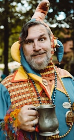 A man dressed in a colorful jester costume, featuring multi-colored fabric and a hat with bells, holds a pewter tankard. He is adorned with beaded necklaces and has a playful expression.