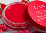 A vibrant red lip balm container with a matching red lid partially opened. The lid has the words 'fresh SUGAR WATERMELON HYDRATING' written in white. The background includes small decorative elements resembling watermelon slices and colorful round beads.
