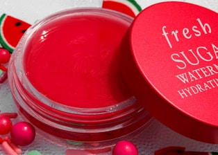 A vibrant red lip balm container with a matching red lid partially opened. The lid has the words 'fresh SUGAR WATERMELON HYDRATING' written in white. The background includes small decorative elements resembling watermelon slices and colorful round beads.