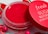 A vibrant red lip balm container with a matching red lid partially opened. The lid has the words 'fresh SUGAR WATERMELON HYDRATING' written in white. The background includes small decorative elements resembling watermelon slices and colorful round beads.