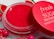 A vibrant red lip balm container with a matching red lid partially opened. The lid has the words 'fresh SUGAR WATERMELON HYDRATING' written in white. The background includes small decorative elements resembling watermelon slices and colorful round beads.