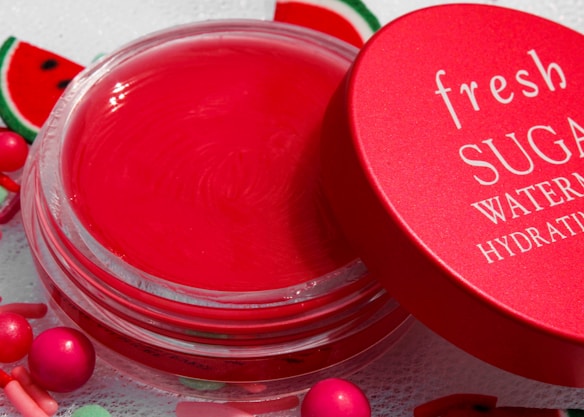A vibrant red lip balm container with a matching red lid partially opened. The lid has the words 'fresh SUGAR WATERMELON HYDRATING' written in white. The background includes small decorative elements resembling watermelon slices and colorful round beads.