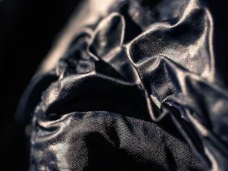 A close-up view of a dark, shiny fabric with visible wrinkles and folds. The fabric has a reflective surface that catches the light, creating a textured and dynamic visual effect.