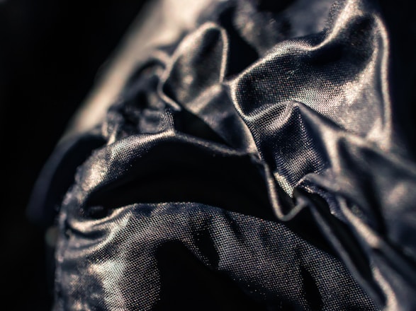 A close-up view of a dark, shiny fabric with visible wrinkles and folds. The fabric has a reflective surface that catches the light, creating a textured and dynamic visual effect.