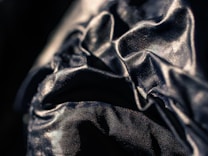 A close-up view of a dark, shiny fabric with visible wrinkles and folds. The fabric has a reflective surface that catches the light, creating a textured and dynamic visual effect.