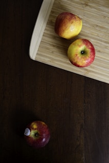 A wooden cutting board is placed on a dark wooden surface, holding two apples with red and yellow hues. A third apple, similar in color, is on the wooden surface, apart from the cutting board. The apple on the surface has a sticker indicating its variety.