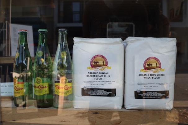 Several bottles of Topo Chico mineral water and two bags of organic flour are displayed on a wooden surface. The bottles have yellow, green, and red labels, and the flour bags are white with detailed labeling.