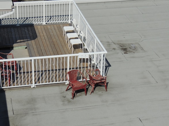 A rooftop deck with wooden flooring is enclosed by white railings. Three white chairs are aligned neatly in the corner of the deck. Outside the enclosed area, two red wicker chairs are stacked together on the flat rooftop surface.