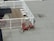 A rooftop deck with wooden flooring is enclosed by white railings. Three white chairs are aligned neatly in the corner of the deck. Outside the enclosed area, two red wicker chairs are stacked together on the flat rooftop surface.