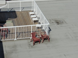 A rooftop deck with wooden flooring is enclosed by white railings. Three white chairs are aligned neatly in the corner of the deck. Outside the enclosed area, two red wicker chairs are stacked together on the flat rooftop surface.