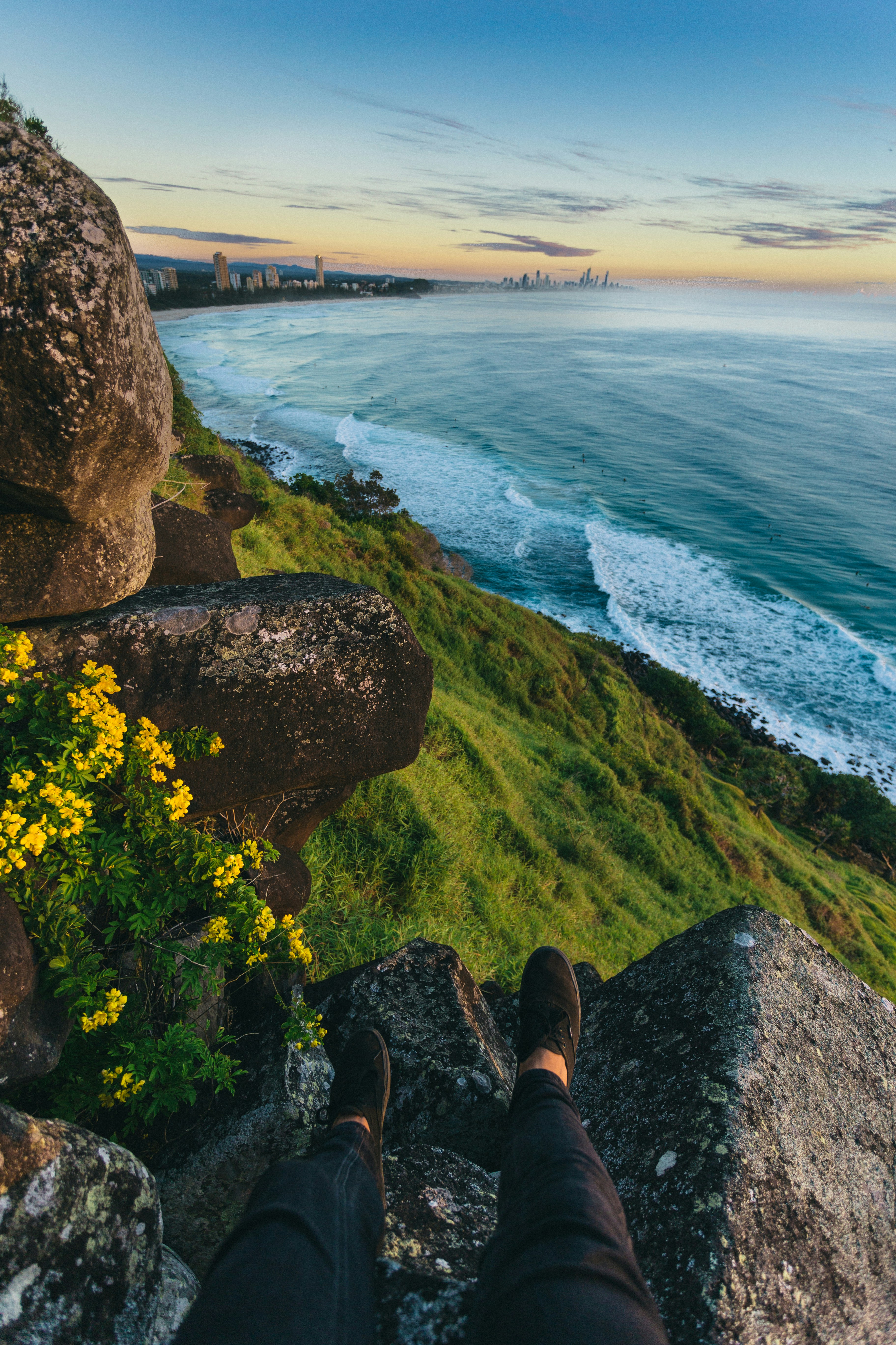 person in black pants and brown shoes sitting on rock near body of water during daytime
