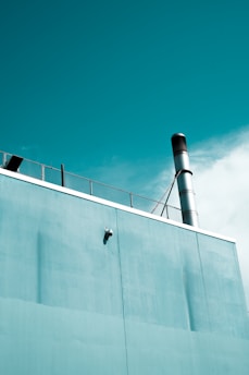A section of a building is captured with a teal-colored wall and a metal chimney extending upwards. A surveillance camera is mounted on the wall and part of a metal fence is visible on the rooftop. The sky is partly cloudy, adding to the serene atmosphere.