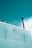 A section of a building is captured with a teal-colored wall and a metal chimney extending upwards. A surveillance camera is mounted on the wall and part of a metal fence is visible on the rooftop. The sky is partly cloudy, adding to the serene atmosphere.
