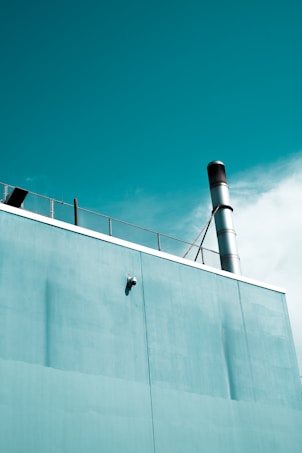 A section of a building is captured with a teal-colored wall and a metal chimney extending upwards. A surveillance camera is mounted on the wall and part of a metal fence is visible on the rooftop. The sky is partly cloudy, adding to the serene atmosphere.