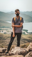 A person stands on rocky terrain with a mountainous landscape in the background. Wearing a backpack and a face mask, they are dressed casually in a t-shirt and pants, suggesting an outdoor adventure.