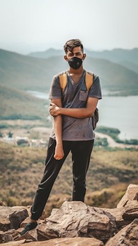 A person stands on rocky terrain with a mountainous landscape in the background. Wearing a backpack and a face mask, they are dressed casually in a t-shirt and pants, suggesting an outdoor adventure.