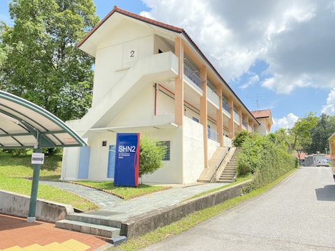 A two-story cream-colored building with red-tiled roof stands under a partly cloudy sky. An external staircase is visible. There's a blue signboard labeled SHN2 near the entrance. The surrounding area has green grass, a paved walkway, and a bus stop shelter on the left. A road runs alongside the building, bordered by greenery and trees.