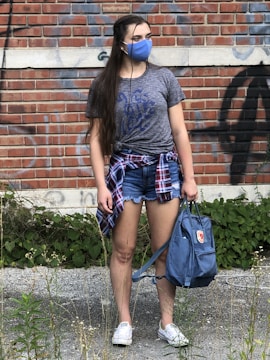 A woman stands outdoors in casual attire, including a gray T-shirt with blue lettering, denim shorts, and white sneakers. She wears a blue face mask and holds a blue bag. A plaid shirt is tied around her waist. The background features a brick wall with graffiti and some green vegetation growing at ground level.