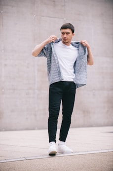 A young man stands confidently, adjusting his striped shirt over a plain white t-shirt. He wears dark pants and white sneakers, set against a minimalistic urban background with a concrete wall.