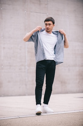 A young man stands confidently, adjusting his striped shirt over a plain white t-shirt. He wears dark pants and white sneakers, set against a minimalistic urban background with a concrete wall.