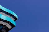 Angular, modern architectural structure with teal-colored panels is positioned against a deep blue, cloudless sky.