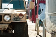A rugged military vehicle is parked on a sunlit street beside a row of chains and bollards. The background contains a mural with abstract graffiti on a wall, and other buildings are visible, contributing to an urban setting.