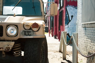 A rugged military vehicle is parked on a sunlit street beside a row of chains and bollards. The background contains a mural with abstract graffiti on a wall, and other buildings are visible, contributing to an urban setting.