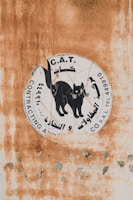 A weathered sign with a graphic of a black cat surrounded by text in Arabic and English. The background features a rusty and peeling surface, indicating age and exposure to the elements.
