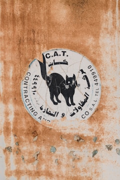 A weathered sign with a graphic of a black cat surrounded by text in Arabic and English. The background features a rusty and peeling surface, indicating age and exposure to the elements.