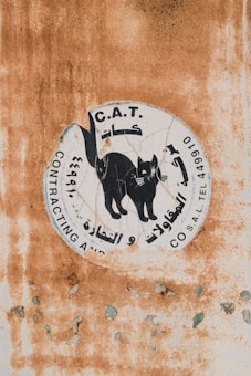A weathered sign with a graphic of a black cat surrounded by text in Arabic and English. The background features a rusty and peeling surface, indicating age and exposure to the elements.