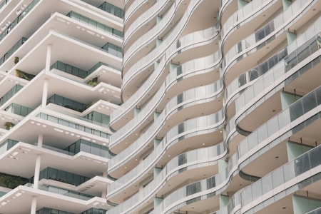 A modern architectural design featuring two multi-story buildings with flowing, wave-like balconies. The structures have a sleek and minimalistic style, with large glass panels and white surfaces. Greenery is visible on some of the balconies, adding a touch of nature to the urban setting.