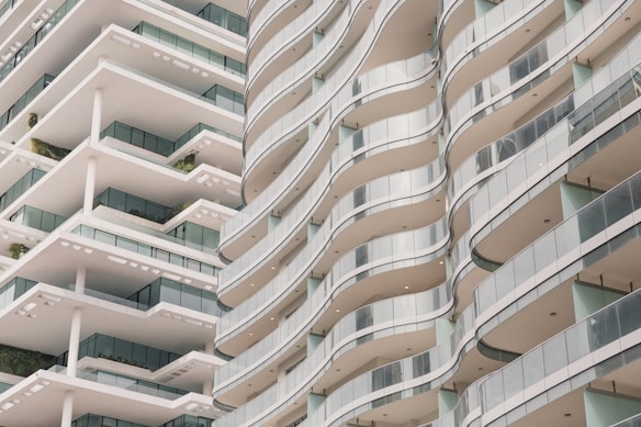 A modern architectural design featuring two multi-story buildings with flowing, wave-like balconies. The structures have a sleek and minimalistic style, with large glass panels and white surfaces. Greenery is visible on some of the balconies, adding a touch of nature to the urban setting.