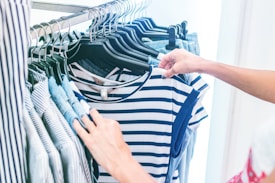 A person is browsing through a rack of clothes, focusing on a striped blue and white shirt. The garments are hung on black hangers and include various striped and solid-colored shirts. The setting appears to be a retail store with soft lighting.