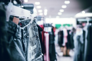 A rack of denim shorts is prominently displayed in the foreground, with several other clothing items blurred in the background. The setting appears to be inside a retail clothing store with bright overhead lighting.
