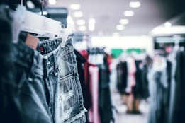 A rack of denim shorts is prominently displayed in the foreground, with several other clothing items blurred in the background. The setting appears to be inside a retail clothing store with bright overhead lighting.