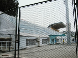 A modern sports stadium with tiered seating and a large, curved roof is seen behind a chain-link fence. The area appears to be an entrance or side of the stadium, featuring concrete structures and some air conditioning units. Two people walk near the entrance, wearing casual clothing.