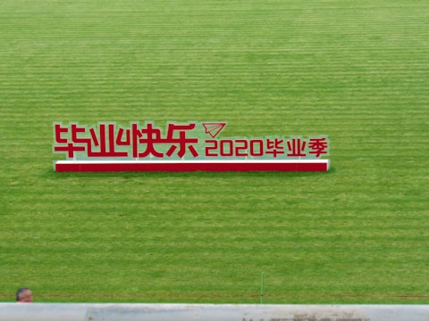 A large, grassy field with evenly mowed stripes. Positioned in the center is a bold sign with red characters and numbers on a white background, reading '2020'. The sign is celebrating a graduation season, indicated by the presence of red text in another language. A person is partially visible in the lower area of the image.