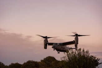 A tiltrotor aircraft is flying in the sky with its propellers in a vertical position. The background features a serene evening sky with a gradient of soft colors. Greenery, including trees and bushes, are visible at the lower part of the image.