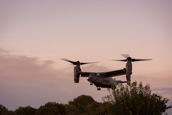 A tiltrotor aircraft is flying in the sky with its propellers in a vertical position. The background features a serene evening sky with a gradient of soft colors. Greenery, including trees and bushes, are visible at the lower part of the image.