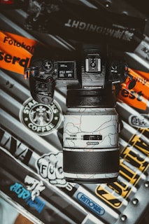 A camera with a distinctive lens is placed on a variety of stickers and decals from brands like Starbucks Coffee, Rimowa, and others. The camera's screen displays settings like ISO 1600 and F5.0. The design on the lens features an abstract pattern with lines and curves on a white background.