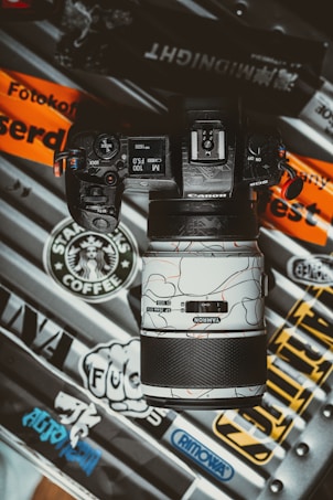 A camera with a distinctive lens is placed on a variety of stickers and decals from brands like Starbucks Coffee, Rimowa, and others. The camera's screen displays settings like ISO 1600 and F5.0. The design on the lens features an abstract pattern with lines and curves on a white background.