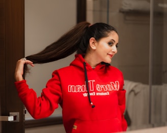 A person is standing indoors, looking at their reflection in a mirror. They are wearing a bright red hooded sweatshirt and have long brown hair styled in a high ponytail. The hand is reaching back to hold the ponytail. The background appears to be a bathroom, with towels visible.